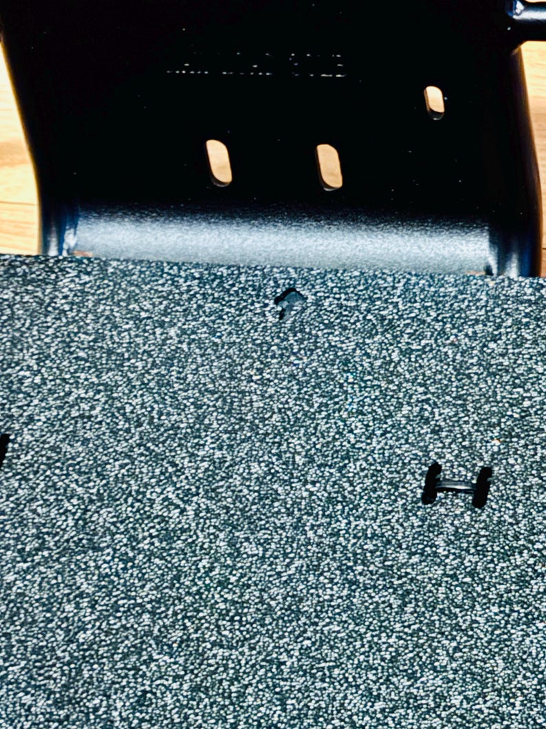 May include: Close-up of a textured, speckled, grey surface with a black metal component. The metal piece has two oval cutouts and a curved shape. The surface has a small, diamond-shaped cutout and a black, H-shaped piece.