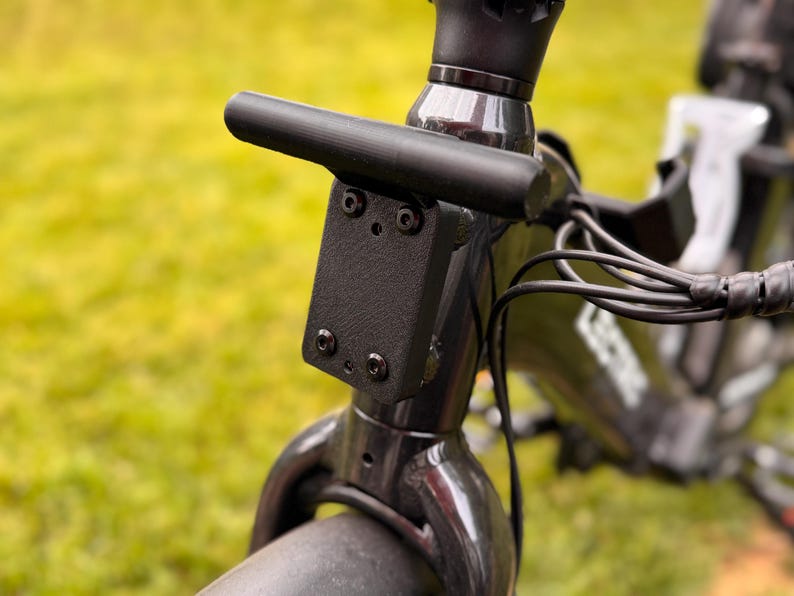 May include: Close-up of a black bicycle handlebar stem with a horizontal bar and a mounting plate. The plate is secured with four screws. Black cables and the front fork are also visible.