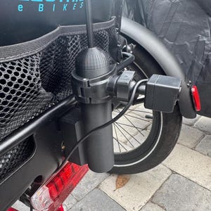 May include: Black accessory mounted on the rear of an electric bicycle. The accessory includes a cylindrical component, a box-shaped component, and a black antenna. The bicycle has a black rear fender, a red frame, and a black storage bag with the text "LECTRIC e BIKES".