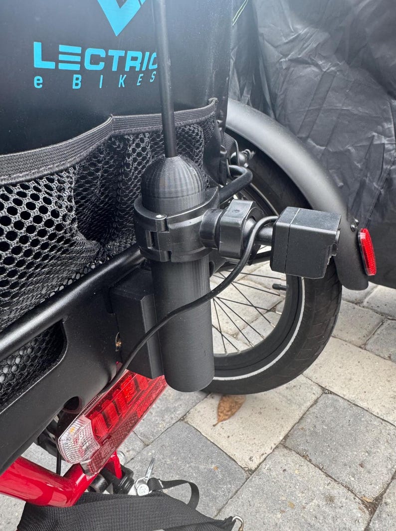 May include: Black accessory mounted on the rear of an electric bicycle. The accessory includes a cylindrical component, a box-shaped component, and a black antenna. The bicycle has a black rear fender, a red frame, and a black storage bag with the text "LECTRIC e BIKES".
