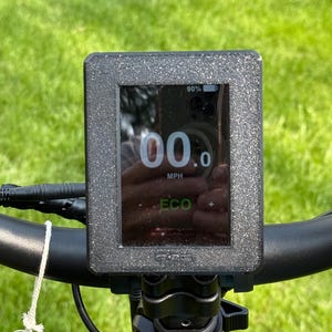 May include: Close-up of an electric bike display showing a speed of 00.0 MPH. The screen displays battery life at 90% and an ECO mode indicator. The display is mounted on black handlebars with a key and warning label visible.