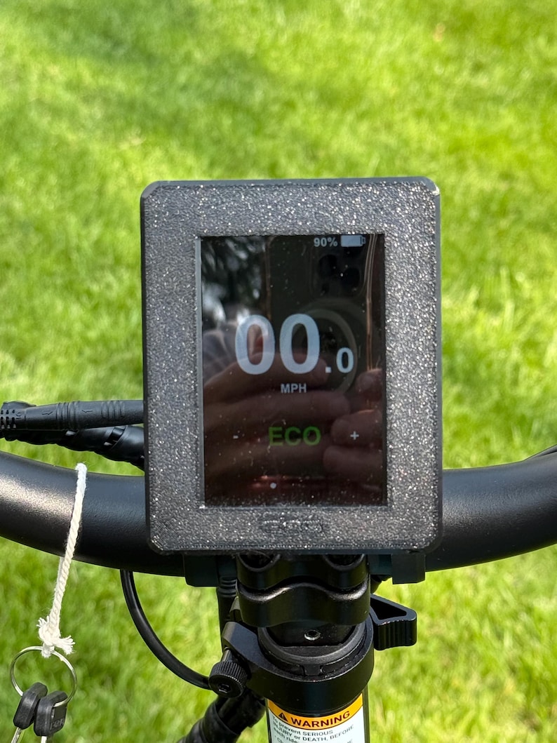 May include: Close-up of an electric bike display showing a speed of 00.0 MPH. The screen displays battery life at 90% and an ECO mode indicator. The display is mounted on black handlebars with a key and warning label visible.