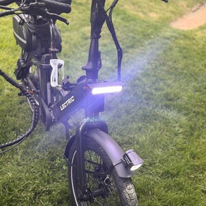 May include: A black Lectric e-bike with a bright white headlight and a front fender. The bike has a black seat, handlebars, and a small bag. The bike is parked on a grassy surface.