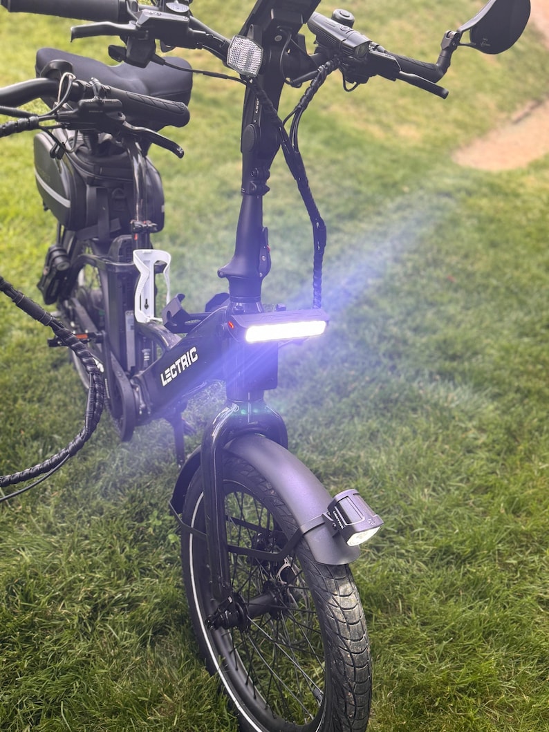 May include: A black Lectric e-bike with a bright white headlight and a front fender. The bike has a black seat, handlebars, and a small bag. The bike is parked on a grassy surface.