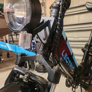 May include: Close-up of a black Lectric e-bike. The image highlights the front section, including the headlight, reflector, and front suspension. The bike frame has blue accents and the word "LECTRIC" is visible.