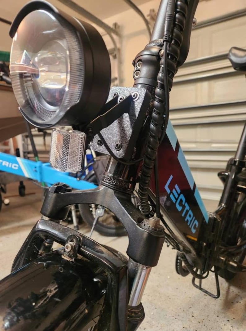 May include: Close-up of a black Lectric e-bike. The image highlights the front section, including the headlight, reflector, and front suspension. The bike frame has blue accents and the word "LECTRIC" is visible.