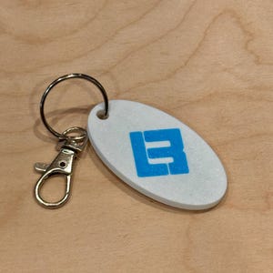 May include: Oval-shaped keychain with a white, glittery surface and a blue letter "B" design. The keychain has a silver-colored clasp and key ring. The background is a light brown wooden surface.