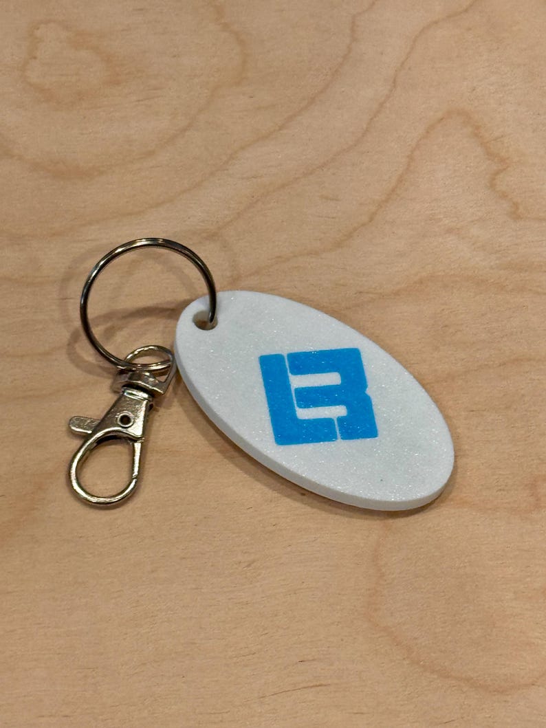 May include: Oval-shaped keychain with a white, glittery surface and a blue letter "B" design. The keychain has a silver-colored clasp and key ring. The background is a light brown wooden surface.