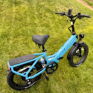 May include: A light blue Lectric ebike with black tires and a black seat is parked on a grassy lawn. The bike has a digital display, a headlight, and a rear rack. The brand name "LECTRIC" is visible on the frame.