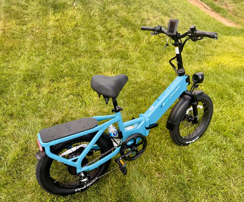 May include: A light blue Lectric ebike with black tires and a black seat is parked on a grassy lawn. The bike has a digital display, a headlight, and a rear rack. The brand name "LECTRIC" is visible on the frame.