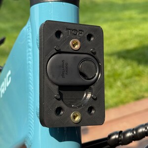May include: A black rectangular mount with the word "TOP" printed on it, attached to a light blue bicycle frame. A black "Galaxy SmartTag2" device is secured within the mount. The bicycle frame has the serial number SNCTC25B167.
