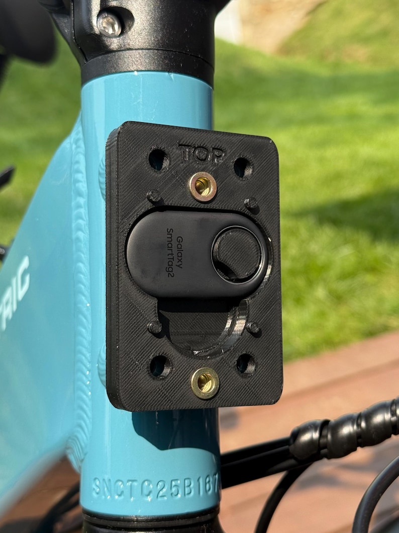 May include: A black rectangular mount with the word "TOP" printed on it, attached to a light blue bicycle frame. A black "Galaxy SmartTag2" device is secured within the mount. The bicycle frame has the serial number SNCTC25B167.