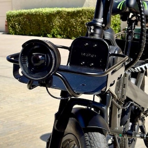 May include: Close-up of a black electric bicycle with a front basket and headlight. The bicycle has a black frame, a round headlight, and a small basket attached to the handlebars. The tires are black and the bike appears to be in good condition.