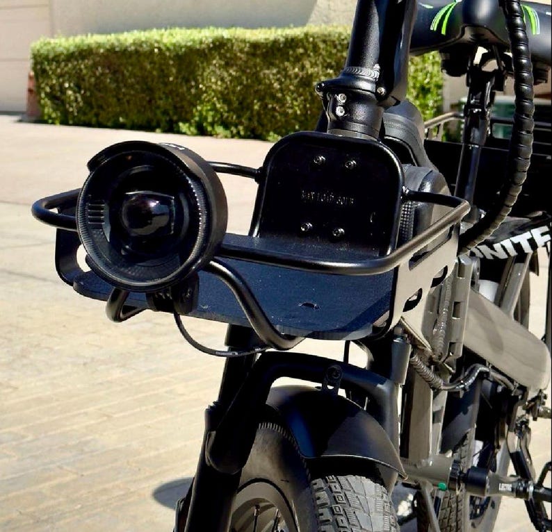 May include: Close-up of a black electric bicycle with a front basket and headlight. The bicycle has a black frame, a round headlight, and a small basket attached to the handlebars. The tires are black and the bike appears to be in good condition.