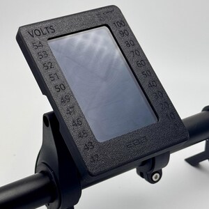 May include: Black electric bike display with a screen showing a voltage reading and a gauge. The display is mounted on a black handlebar with a green and black wire.