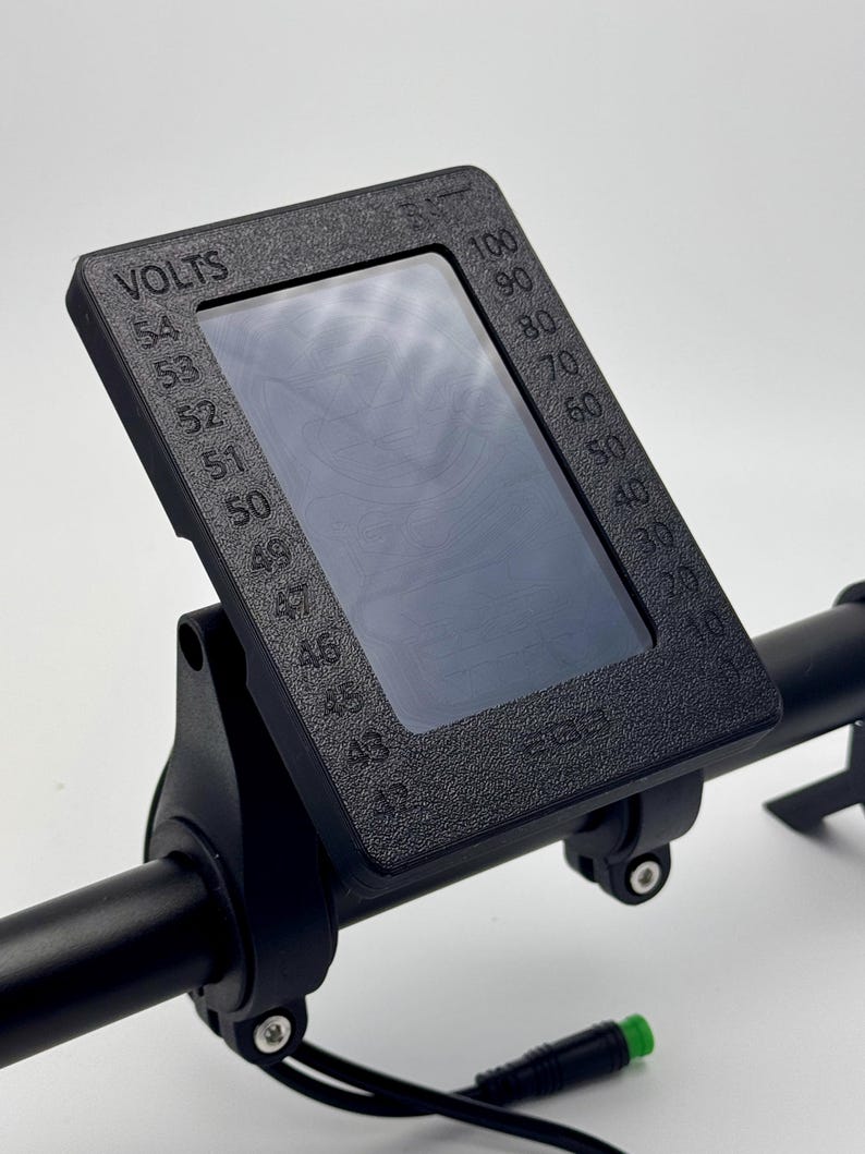May include: Black electric bike display with a screen showing a voltage reading and a gauge. The display is mounted on a black handlebar with a green and black wire.
