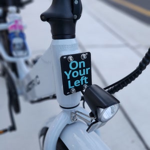 May include: A white electric bicycle with a black handlebar and a black light. The bicycle has a sign that says "On Your Left" in blue text.