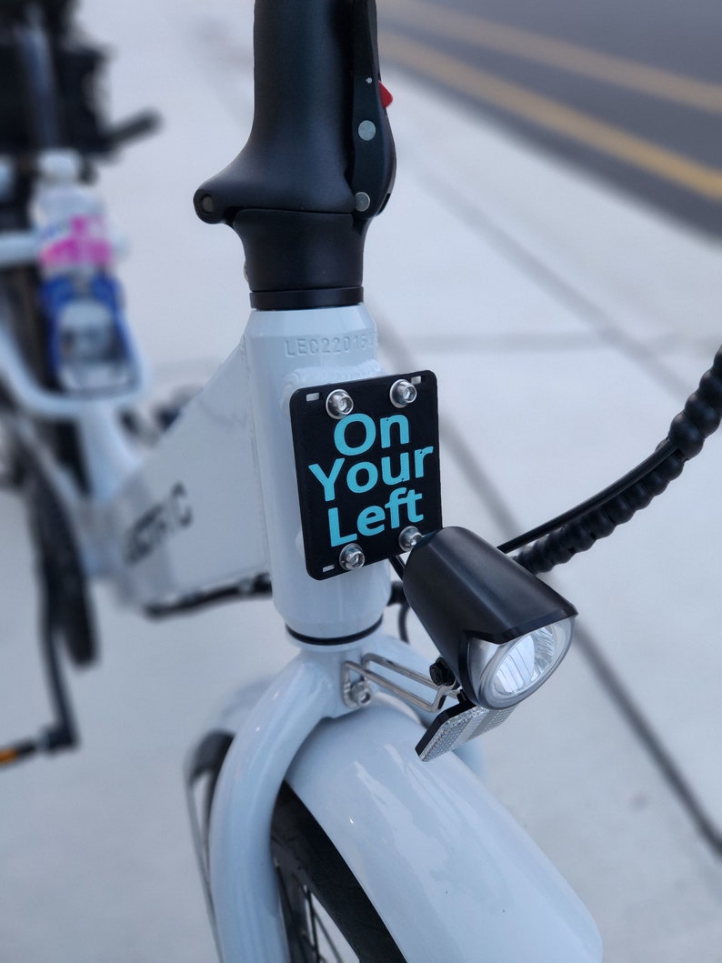 May include: A white electric bicycle with a black handlebar and a black light. The bicycle has a sign that says "On Your Left" in blue text.