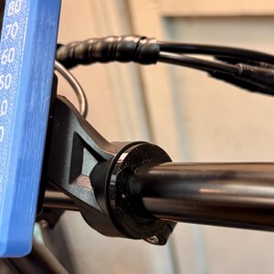 Op de afbeelding: Close-up van een zwart stuur van een fiets met een zwarte klem eraan bevestigd. De klem heeft een schroef aan de onderkant.