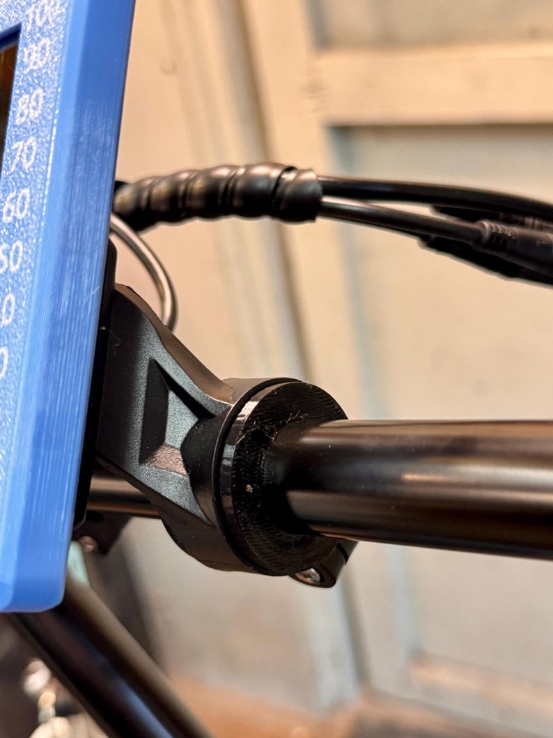 Op de afbeelding: Close-up van een zwart stuur van een fiets met een zwarte klem eraan bevestigd. De klem heeft een schroef aan de onderkant.