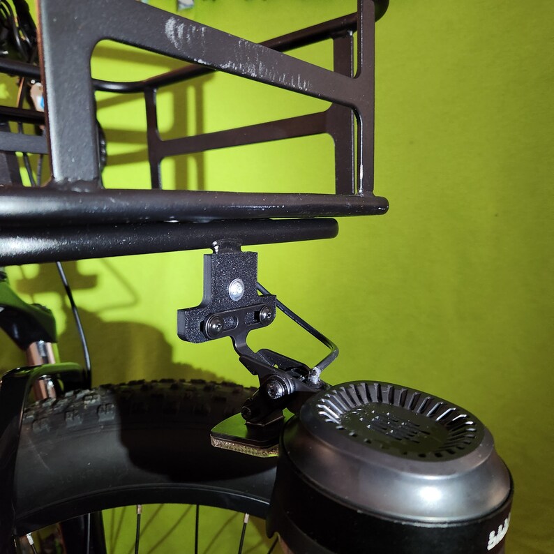 May include: A black metal bike rack with a black metal bracket attached to the bottom. The bracket is attached to a black electric bike motor.