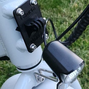 May include: A white electric bicycle with a black headlight and a black camera mount attached to the handlebars. The camera mount has the word "Camera" printed on it.