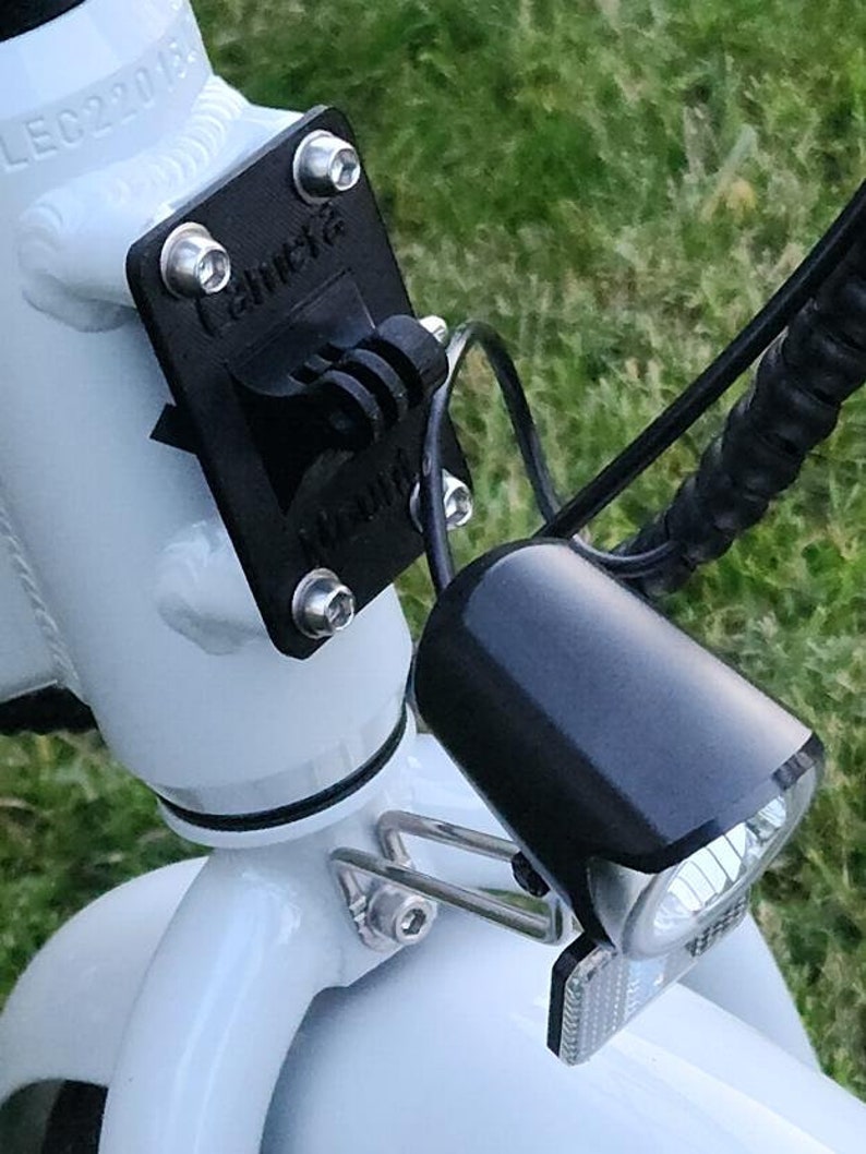 May include: A white electric bicycle with a black headlight and a black camera mount attached to the handlebars. The camera mount has the word "Camera" printed on it.