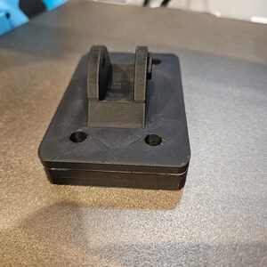 May include: A black, rectangular 3D-printed object with a raised bracket. The object has two circular holes and a textured surface. The bracket has two rounded sections. The item appears to be a component or part.