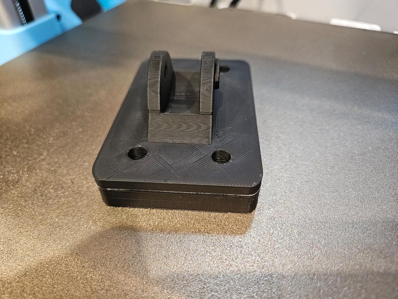 May include: A black, rectangular 3D-printed object with a raised bracket. The object has two circular holes and a textured surface. The bracket has two rounded sections. The item appears to be a component or part.