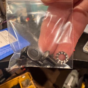 May include: A clear plastic bag containing a variety of small black metal screws and washers. The bag is held in a person's hand.
