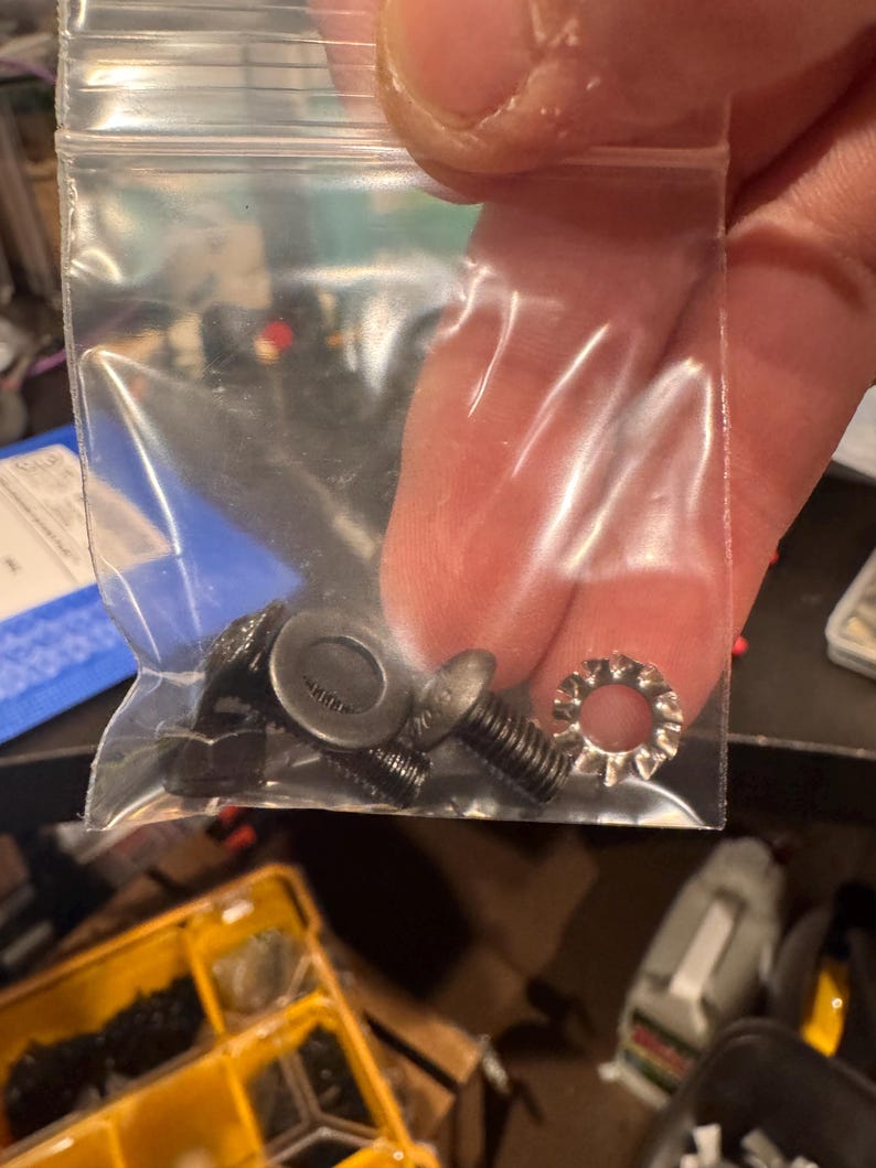 May include: A clear plastic bag containing a variety of small black metal screws and washers. The bag is held in a person's hand.