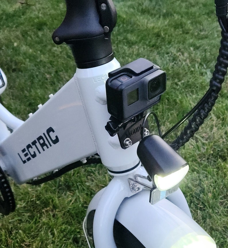 May include: A white electric bike with a black camera mounted on the handlebars. The bike has a black handlebar, black seat, and black tires. The camera is a GoPro and is mounted on a black mount. The bike has a black light on the front.