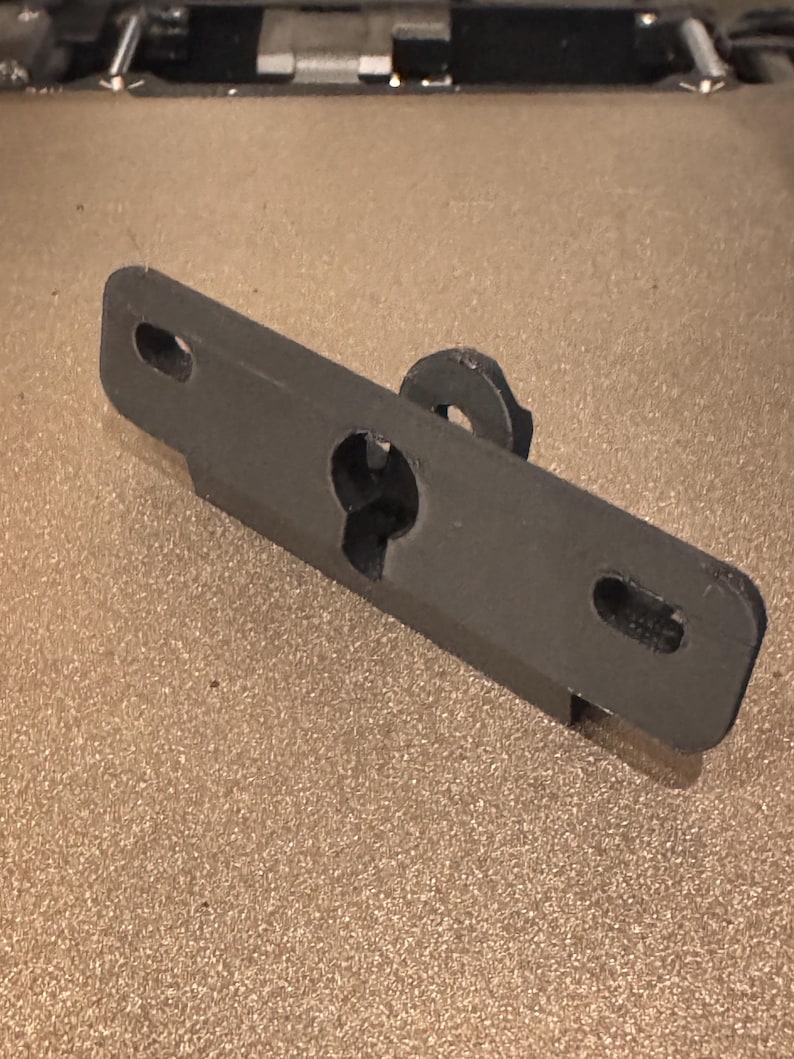 May include: A black plastic bracket with a hole in the center and two smaller holes on either side. The bracket has a small, round knob on the top.