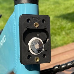 May include: Black 3D-printed mount with an Apple AirTag installed on a light blue bicycle frame. The mount is labeled "TOP" and secured with brass-colored screws. The bicycle frame has the word "ELECTRIC" printed on it.
