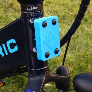 May include: Close-up of a black electric bicycle with a blue plastic plate attached to the frame. The plate has a logo and five black screws. The bicycle has a black handlebar and a black seat.