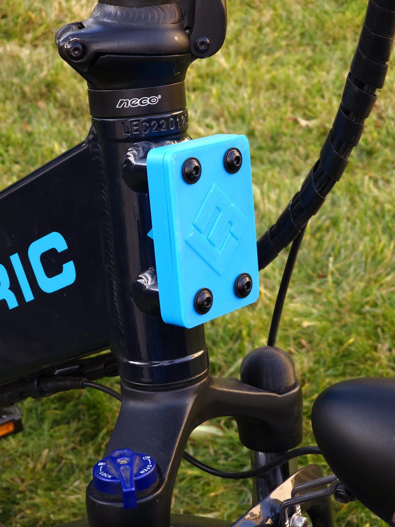 May include: Close-up of a black electric bicycle with a blue plastic plate attached to the frame. The plate has a logo and five black screws. The bicycle has a black handlebar and a black seat.