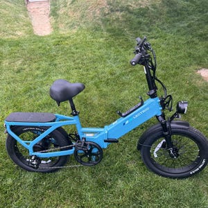 May include: A light blue electric bicycle with black tires and seat is parked on a grassy lawn. The bike has a black handlebar, a headlight, and a rear rack. The brand name "LECTRIC" is visible on the frame.