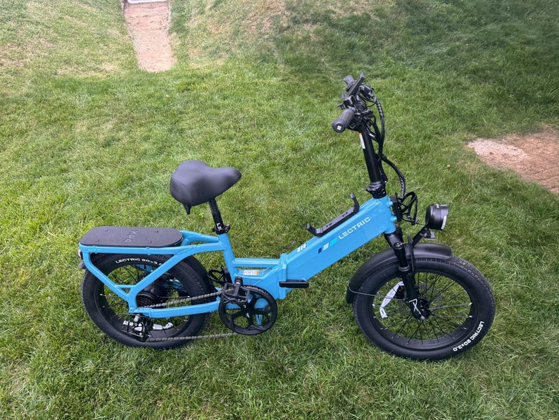 May include: A light blue electric bicycle with black tires and seat is parked on a grassy lawn. The bike has a black handlebar, a headlight, and a rear rack. The brand name "LECTRIC" is visible on the frame.