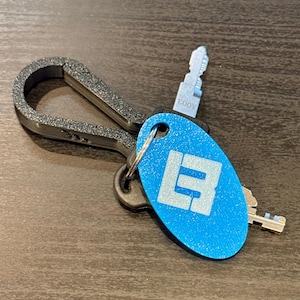 May include: A set of keys attached to a black carabiner clip. The keys include a blue oval tag with a white logo and a silver key with the text "A003". The keys are on a dark brown surface.