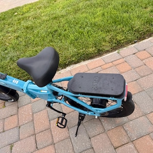 May include: A light blue electric bicycle with a black seat and a black rear cargo rack. The bike has black tires and pedals. The brand name "Xp" is visible on the cargo rack.