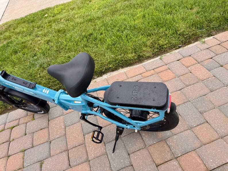 May include: A light blue electric bicycle with a black seat and a black rear cargo rack. The bike has black tires and pedals. The brand name "Xp" is visible on the cargo rack.