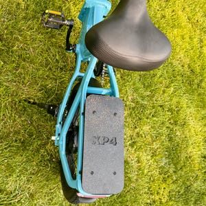 May include: A light blue folding bicycle with a black seat and a black footrest. The footrest has the text "XP4" printed on it. The bicycle is on a grassy surface.