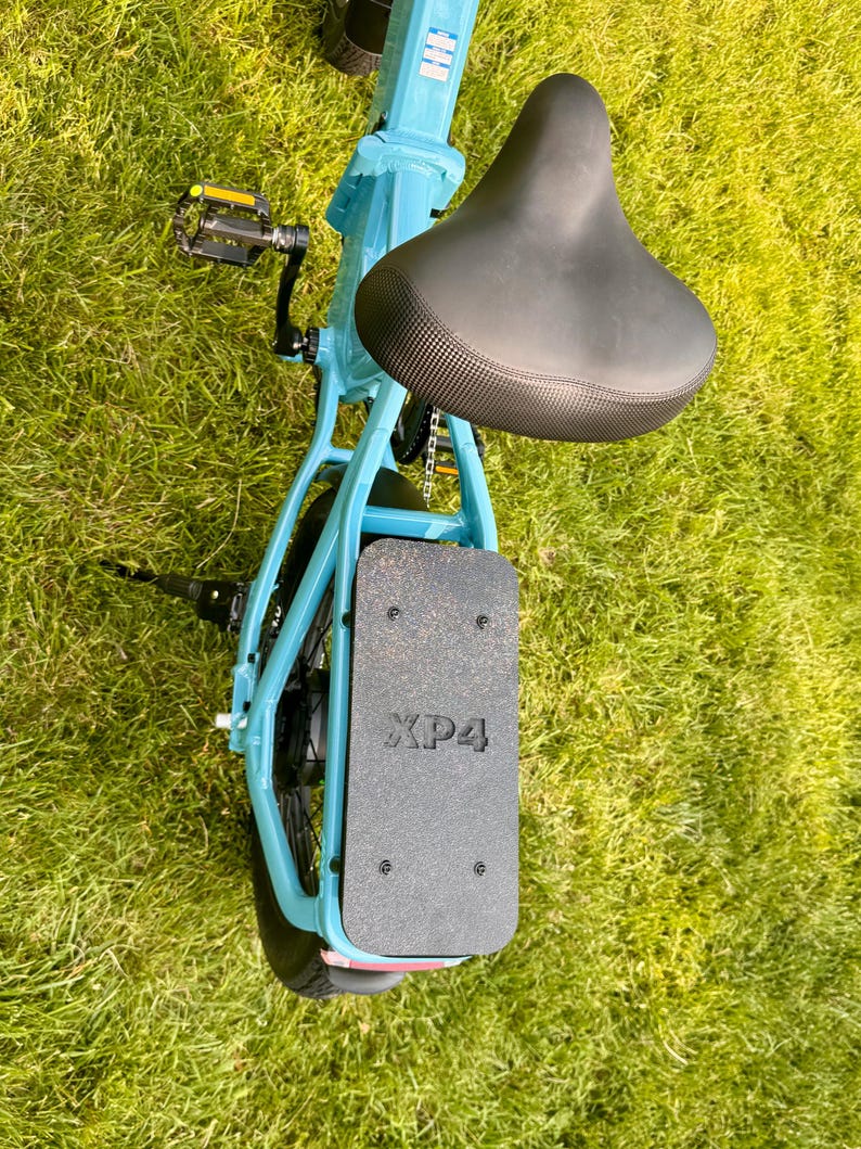 May include: A light blue folding bicycle with a black seat and a black footrest. The footrest has the text "XP4" printed on it. The bicycle is on a grassy surface.
