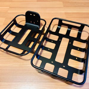 May include: Two black metal bicycle baskets. One has a mounting bracket, the other is rectangular. The baskets are made of connected bars, designed to hold items on a bicycle. The baskets are made of metal.