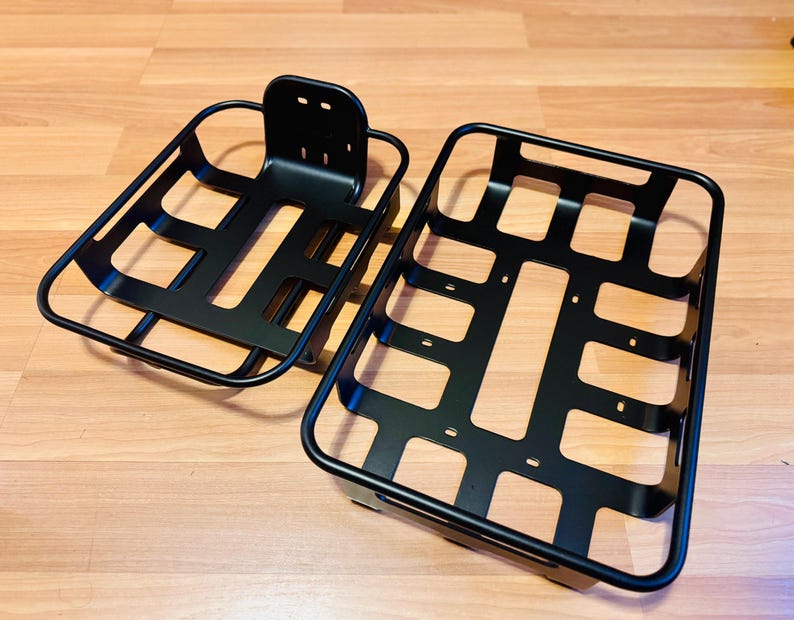 May include: Two black metal bicycle baskets. One has a mounting bracket, the other is rectangular. The baskets are made of connected bars, designed to hold items on a bicycle. The baskets are made of metal.