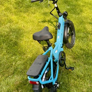 May include: A light blue Lectric XP 4 electric bicycle with black tires, seat, and handlebars. The bike has a digital display, a rear rack, and a red taillight. It is positioned on a grassy surface.