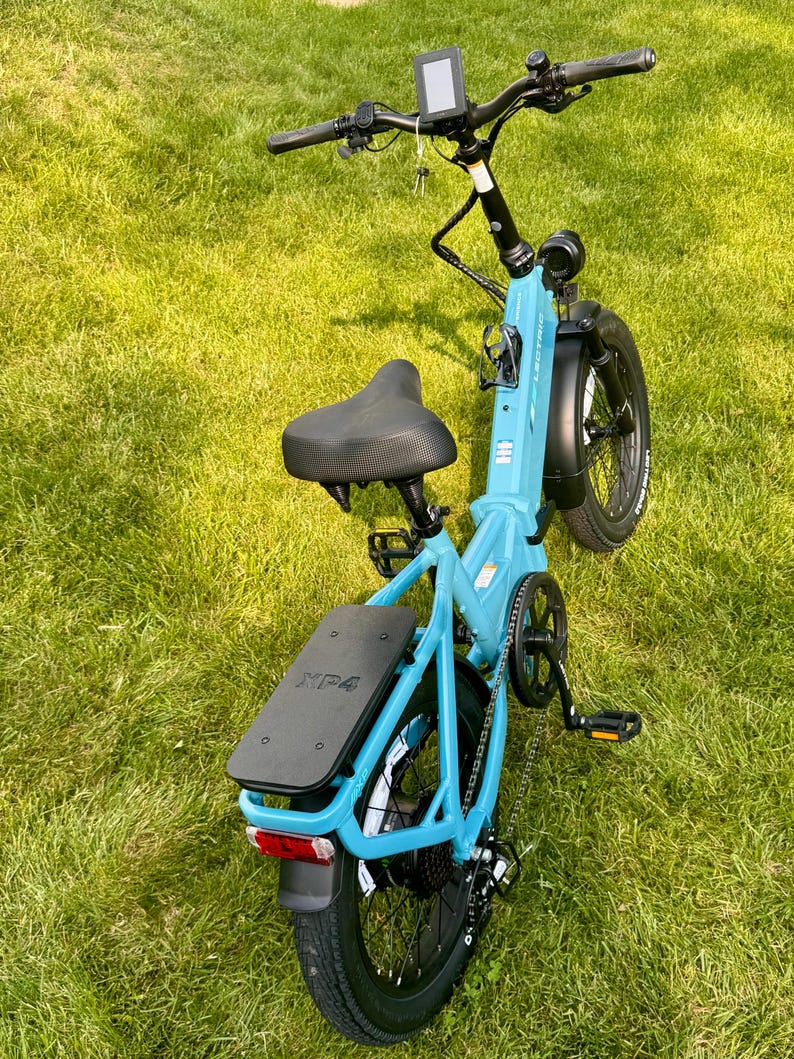 May include: A light blue Lectric XP 4 electric bicycle with black tires, seat, and handlebars. The bike has a digital display, a rear rack, and a red taillight. It is positioned on a grassy surface.
