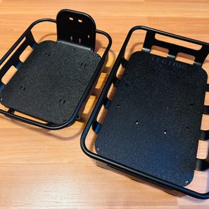 May include: Two black metal bicycle baskets. One has a back support with mounting holes. The other has the word "LECTRIC" printed on the base. Both have a rectangular shape with a textured base.