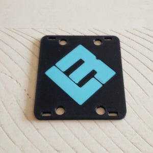 May include: Black plastic plate with four holes and a blue logo in the center. The logo is a stylized letter "L" with a smaller "L" inside.