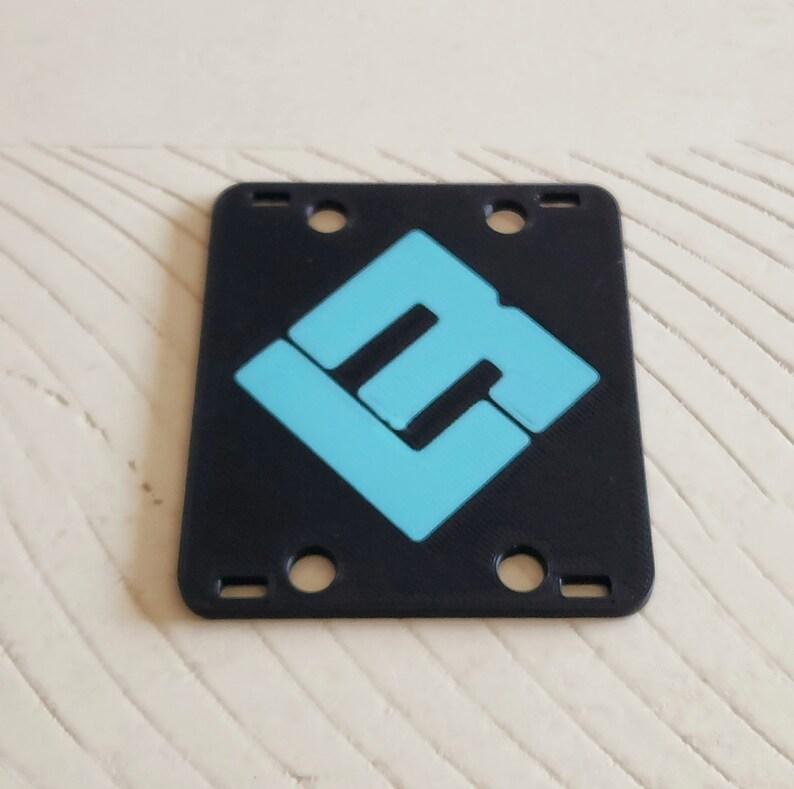May include: Black plastic plate with four holes and a blue logo in the center. The logo is a stylized letter "L" with a smaller "L" inside.